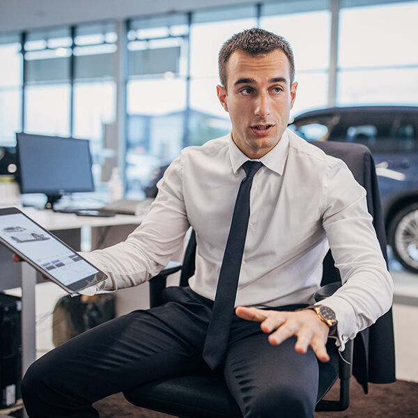 Young salesman in shirt and tie holds a tablet device in his hand. The tablet screen shows a car sales website