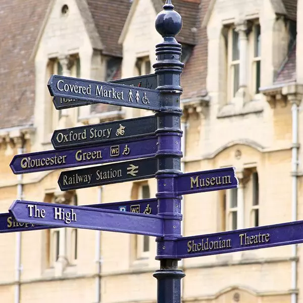 "Blue street sign in Oxford, England with many directions in front of classing Oxford architecture."